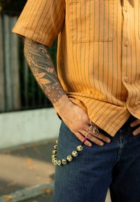 Mann in orange gestreiftem Hemd und blauen Jeans, mit einem Würfel-Schlüsselanhänger, der an der Gürtelschlaufe hängt, und einem detaillierten Armtattoo, das sichtbar ist.