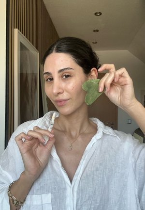 Bouteille de sérum en verre clair dans la main, portant une chemise blanche ample. Tient un outil facial en jade vert en forme de cœur. Éclairage naturel.