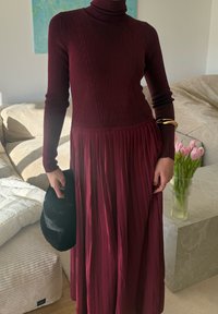 Personne portant une robe longue en turtleneck côtelée bordeaux, tenant un chapeau noir, debout près d'un vase de tulipes roses sur une table en marbre.