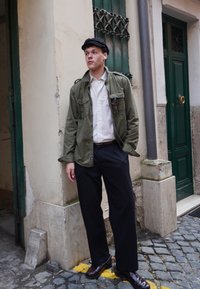 Olivgrüne Utility-Jacke über einem weißen Hemd, schwarzer Hose und dunklen Schuhen. Der Schauspieler steht neben einer strukturierten Wand und grünen Türen.