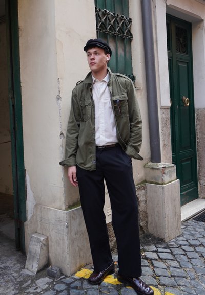 Chaqueta de utilidad verde oliva sobre una camisa blanca, pantalones negros y zapatos oscuros. El actor está de pie cerca de una pared texturizada y puertas verdes.