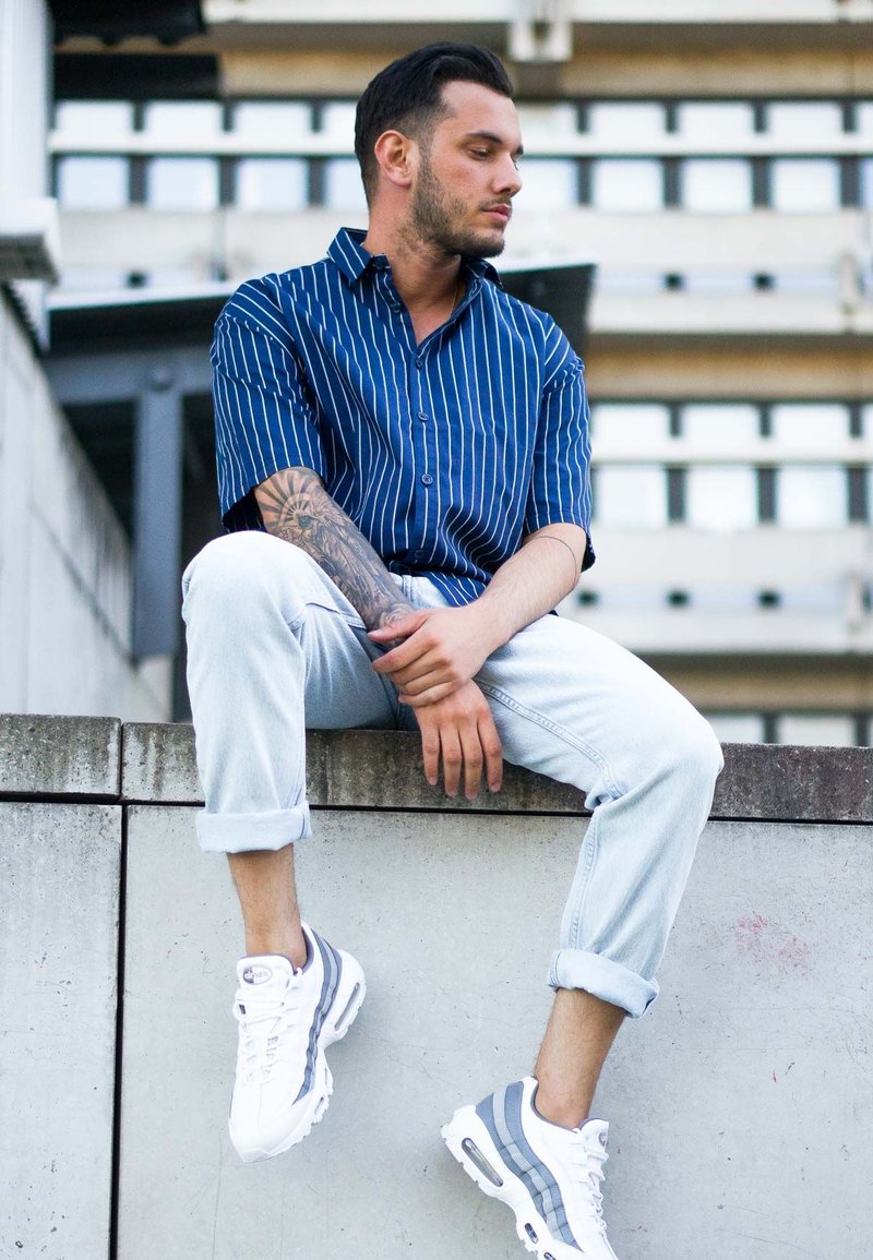 Blau gestreifte Kurzarmbluse, hellblaue hochgekrempelte Jeans und weiße Sneakers mit grauen Akzenten. Stehend auf einer Betonkante.