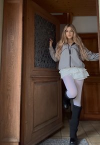 Jachetă gri cu fermoar, fustă albă cu volane și colanți albi. Șosete negre până la genunchi și cizme. Ușa din lemn are un design ornamentat.