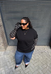 Femeie îmbrăcată cu mănuși negre cu model de lună în formă de semicerc, bluză neagră, blugi albaștri, pantofi negri cu vârful ascuțit, ochelari de soare mari și un colier cu pandantiv.