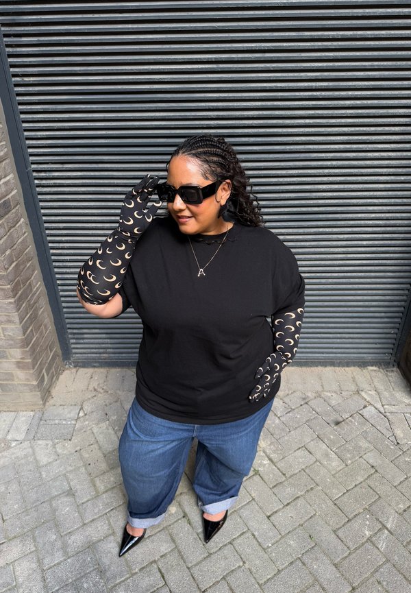 Frau trägt schwarze Handschuhe mit Halbmondmuster, ein schwarzes Oberteil, blaue Jeans, schwarze spitze Schuhe, große Sonnenbrille und eine Anhänger-Neklace.