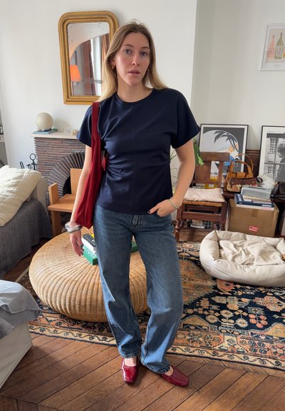 Joven mujer con camiseta azul marino, jeans azules y zapatos rojos, de pie en una sala de estar acogedora sosteniendo un bolso rojo de hombro con una mano en el bolsillo.