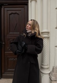 Tumšs vilnas virsjaka ar lielu apkakli, aizpogāta ar vienu pogu, kopā ar melnām ādas cimdiņām un gaišu augstu apkakli zem tā.