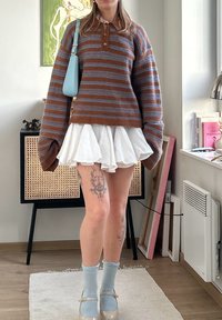 Brun og blåstribet strikket sweater, hvid udvidet nederdel, blå taske, lyseblå sokker, klare sko. Synlig tatovering på benet. Minimalistisk baggrund.