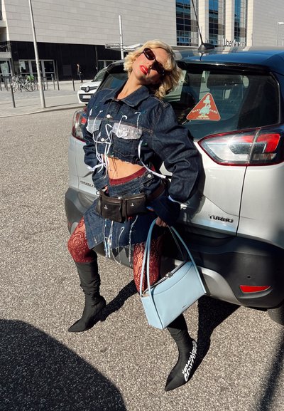 Mujer con chaqueta corta de denim y falda, medias rojas con estampado, botas negras, gafas de sol, apoyada en un coche plateado, sosteniendo un bolso azul claro en la calle.