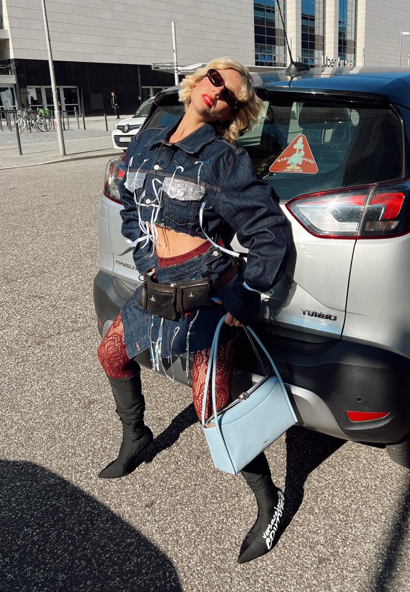 Femme en veste courte en jean et jupe, collants rouges à motifs, bottes noires, lunettes de soleil, appuyée sur une voiture argentée, tenant un sac à main bleu clair dans la rue.