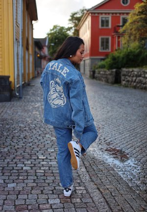 Mujer con chaqueta de mezclilla con logo de Yale estira la pierna en una calle adoquinada con edificios coloridos y vegetación al fondo.