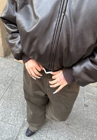 Άτομο που φοράει καφέ δερμάτινο μπουφάν και σκούρο παντελόνι, με χέρια που έχουν μακριά λευκά νύχια και κόκκινα σχέδια με μελάνι, στέκεται σε πλακόστρωτο πεζοδρόμιο.