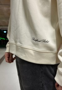 Krém színű pulóver texturált anyagból, bordázott szegéllyel, alján fekete hímzett "Redefined Rebel" felirattal.