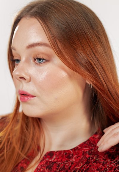 Mujer con cabello largo y rojo, piel clara, ojos azules, vestida con una blusa roja con estampado, mirando ligeramente hacia la izquierda con la mano cerca del cabello.