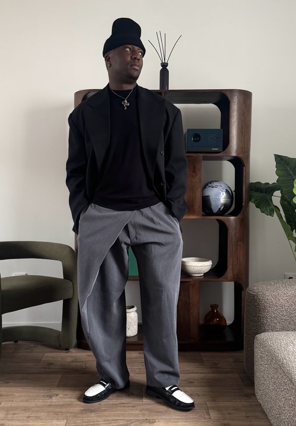 Muž v čiernej čiapke, čiernej bunde a sivých širokých nohaviciach stojí vo vnútri s rukami v vreckách blízko drevenej police a zelenej stoličky.