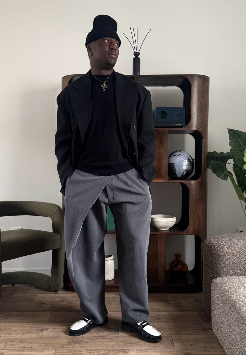 Άνδρας με μαύρο σκουφί, μαύρο μπουφάν και γκρι φαρδιά παντελόνια στέκεται μέσα σε δωμάτιο με τα χέρια στις τσέπες κοντά σε ξύλινο ράφι και πράσινη καρέκλα.