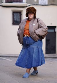 Άτομο που φοράει καφέ γούνινο καπέλο, γυαλιά, ριγέ πουκάμισο, μπεζ μπουφάν, μπλε τζιν φούστα και μπλε παπούτσια slip-on, στέκεται σε πεζοδρόμιο της πόλης.