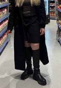 Cappotto lungo nero sopra un top aderente, abbinato a pantaloni corti neri e stivali neri alti fino al ginocchio con suole spesse. Scaffali del negozio sullo sfondo.
