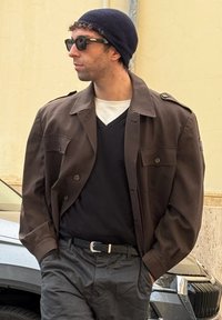 Homme portant un bonnet noir, des lunettes de soleil fonc ées, une veste marron, un pull à col en V noir, une chemise blanche et un pantalon gris, debout les mains dans les poches.