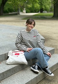 Ριγέ λευκό και μαύρο πουκάμισο με μακριά μανίκια, μπλε τζιν, μαύρα Converse παπούτσια και μία κρεμ τσάντα με κόκκινες γραφικές παραστάσεις.