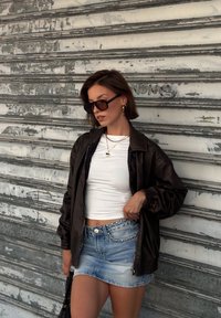 Frau in schwarzer Jacke, weißem Crop-Top, besetztem Denim-Rock, Sonnenbrille und goldener Halskette steht vor einer verwitterten Metallsch shutter.