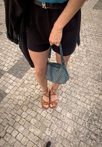 Tīrkīza rokassoma ar kniedēm, ar rāvējslēdzēju un īsu rokturi, apvienota ar brūnām siksniņām sandalēm, melniem šortiem un tumšu jaku. Olevētu bruģa fons.