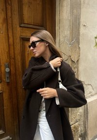 Hnědý vlněný kabát s širokým límcem, bílý top, bílé kalhoty a malá bílá kabelka. Černé sluneční brýle; dřevěné dveře na pozadí.