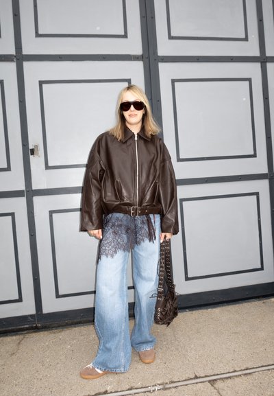 Persona con chaqueta de cuero marrón extragrande, vaqueros de pierna ancha azul claro con detalle de encaje negro, zapatos marrones, gafas de sol y bolso negro en la mano.