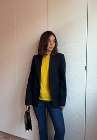 Νεαρή γυναίκα με καστανά μαλλιά μέχρι τους ώμους, που φοράει κίτρινη μπλούζα, μαύρο σακάκι και μπλε τζιν, κρατώντας μια μαύρη τσάντα, όρθια μέσα σε εσωτερικό χώρο.