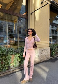 Różowy dopasowany dwuczęściowy strój z długim rękawem na zamek i rozkloszowanymi spodniami. Modelka ma na sobie brązowe obuwie i trzyma małą brązową torebkę.