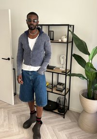 Mand iført en blå ribbestrikket zip-up sweater over en hvid t-shirt, denim shorts og brune ankelstøvler, stående ved siden af en plante og en reol.