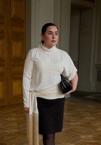 Hvid bluse med høj hals og ballonærmer, kombineret med et sort nederdel. Sort clutch-taske og et lyst tørklæde med frynser.