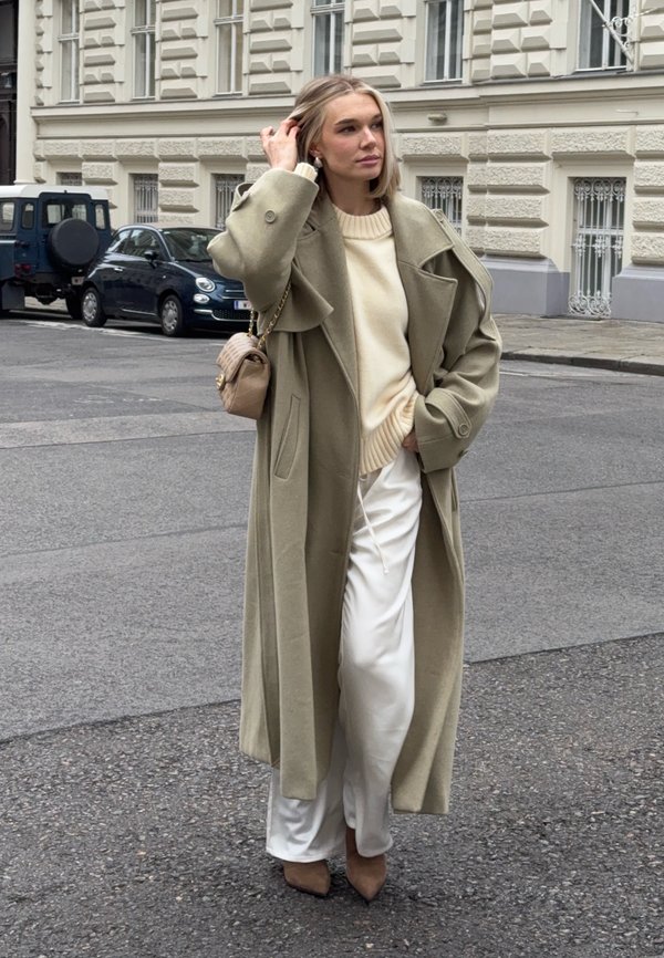 Blond kvinne står i gaten, iført en lang beige frakk, kremfarget genser, hvite bukser, brune støvler, og holder en beige veske.