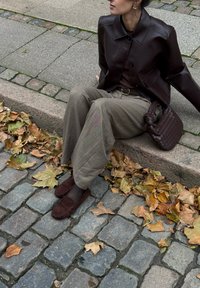 Braune Lederjacke, beige karierte weitbeinige Hose, braune Loafers und eine strukturierte dunkle Tasche, sitzend auf einem Steinweg mit Herbstblättern.