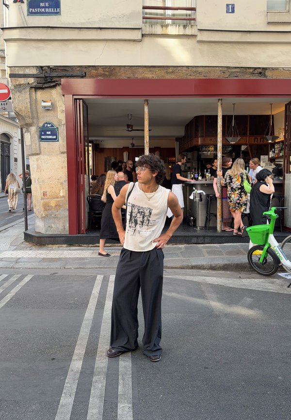 Jeune personne aux cheveux bouclés et portant un débardeur debout dans une rue devant un café ouvert avec des gens à l'intérieur et un vélo de location vert à proximité.