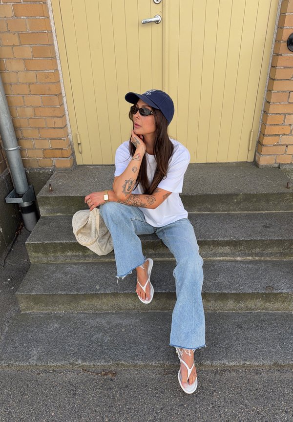 Femme aux longs cheveux et tatouages portant des lunettes de soleil, une casquette, un t-shirt blanc, un jean et des tongs assise sur des marches en pierre à l'extérieur près d'une porte jaune.