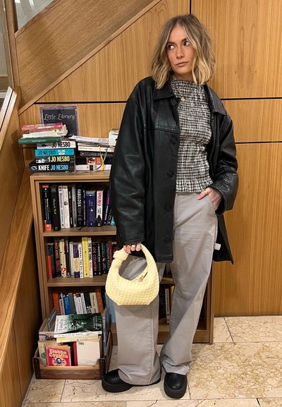 Abrigo oversized de cuero negro, blusa a cuadros, pantalones grises y un bolso tejido amarillo. Texturas suaves, diseño contemporáneo y colores neutros.