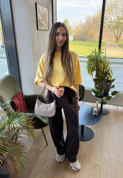 Joven mujer con suéter amarillo y pantalones negros sosteniendo una taza de café y un bolso blanco dentro de una cafetería con plantas y suelo de madera.
