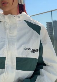 Giacca sportiva bianca con una striscia verde, logo sul petto, chiusura con zip e tessuto texturizzato. Sullo sfondo si vedono edifici e cielo blu.