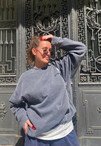 Sweatshirt cinza oversized com ombros caídos, sobre uma camisa branca. Óculos de sol redondos, cabelo longo e esmalte vermelho nas unhas. Fundo metálico decorativo.