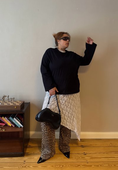 Mujer con top negro, falda blanca de encaje asimétrica, pantalones con estampado de leopardo, zapatos negros puntiagudos, gafas de sol, sujetando un bolso negro junto a una estantería.