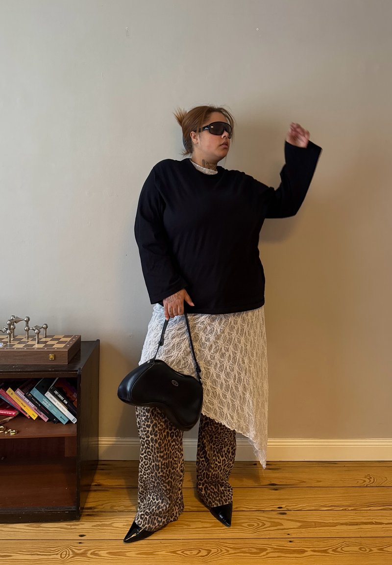 Donna con top nero, gonna asimmetrica in pizzo bianco, pantaloni fantasia leopardata, scarpe nere con punta, occhiali da sole, che tiene una borsa nera vicino a una libreria.