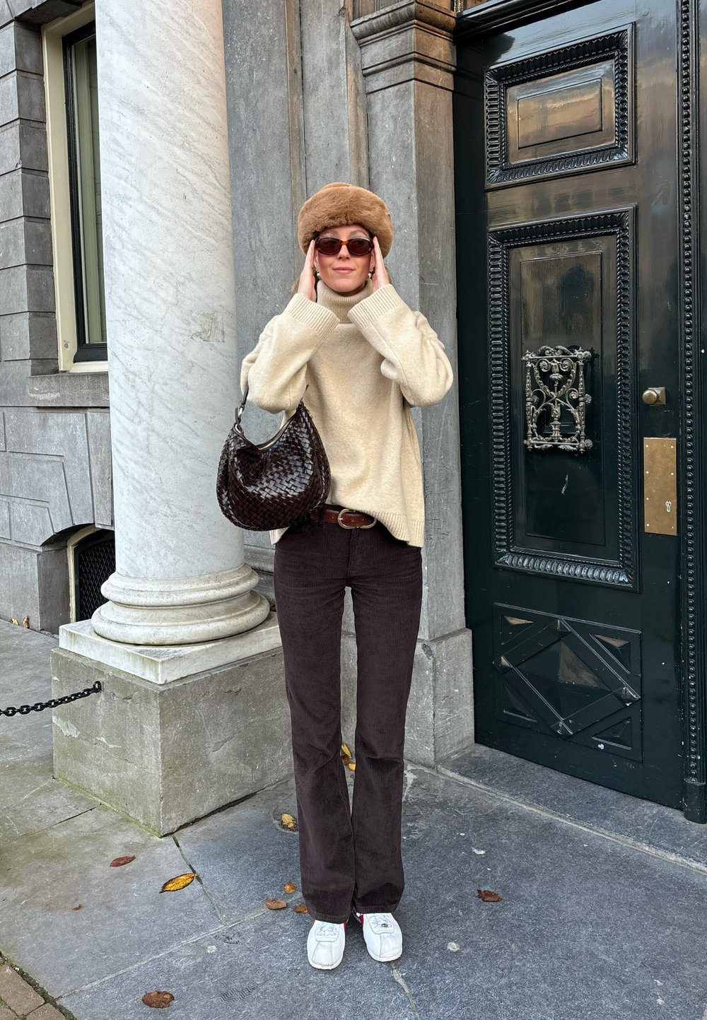 Beiger gestrickter Rollkragenpullover, braune Cordhosen mit weitem Bein, braune gewebte Handtasche, Sonnenbrille und brauner Pelzmütze. Steht neben einer grünen Tür.