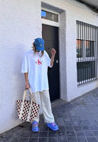 Hvid oversized t-shirt med rødt logo, stribede lyse bukser, blå cap, mønstret tote bag og blå klipklappere med farverige accenter.