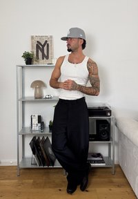 Canotta bianca, pantaloni neri e un cappello grigio. Tatuaggi visibili sulle braccia. Sullo sfondo, uno scaffale con una lampada, piante e vari oggetti.