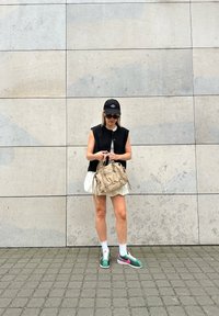 Svart ermeløs bluse over en hvit kjole, beige veske med frynser, grønne joggesko med rosa detaljer, hvite sokker; står mot en teksturert vegg.