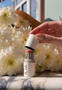 Ilia κρέμα ρουζ σε μορφή στικ με λευκό κυλινδρικό σώμα, ασημί λεπτομέρειες και απαλό ροζ καπάκι, τοποθετημένο μπροστά σε λευκά λουλούδια πάνω σε μαρμάρινη επιφάνεια.