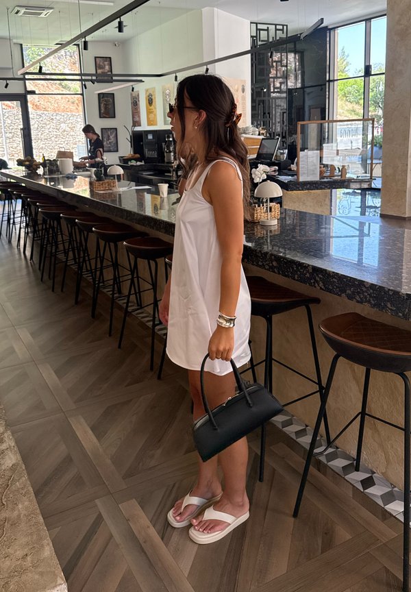 Mujer con vestido blanco y sandalias, sosteniendo un bolso negro, está de pie dentro de una cafetería moderna cerca de taburetes de bar y un mostrador de mármol.
