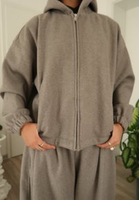 Sweat à capuche gris avec fermeture éclair, coupe décontractée, manches longues avec poignets élastiques et deux poches avant. Assorti à un pantalon gris assorti.