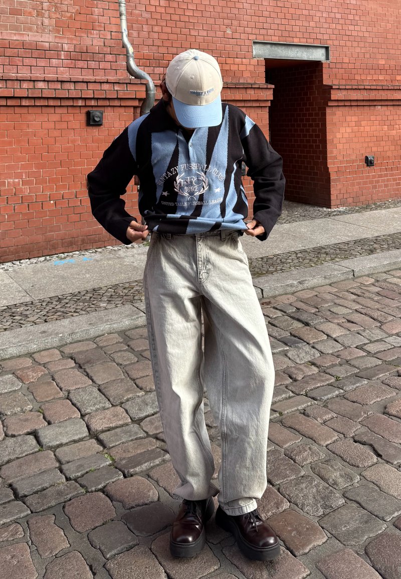 Black and light blue striped long-sleeve shirt with a graphic, light-colored baggy pants, and dark shoes. Light cap completes the look.